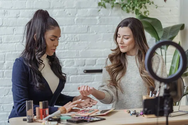 Young positive multiethnic female bloggers testing makeup products while sitting at table and recording vlog on smartphone placed on tripod with ring light. What best creator outreach tools involves in practice