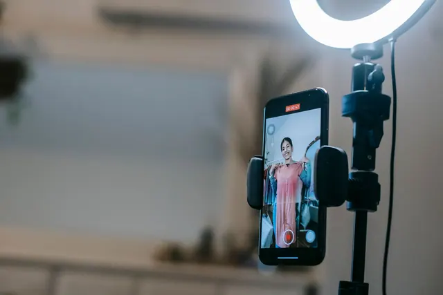 Anonymous female blogger standing in room and shooting vlog on smartphone with ring lamp while demonstrating pink dress in hands. Content approvals: a source-based guide for 2027